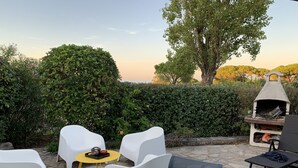 Un espacio al aire libre con sillas blancas y una mesa, un horno de pizza, y vistas a árboles y colinas.