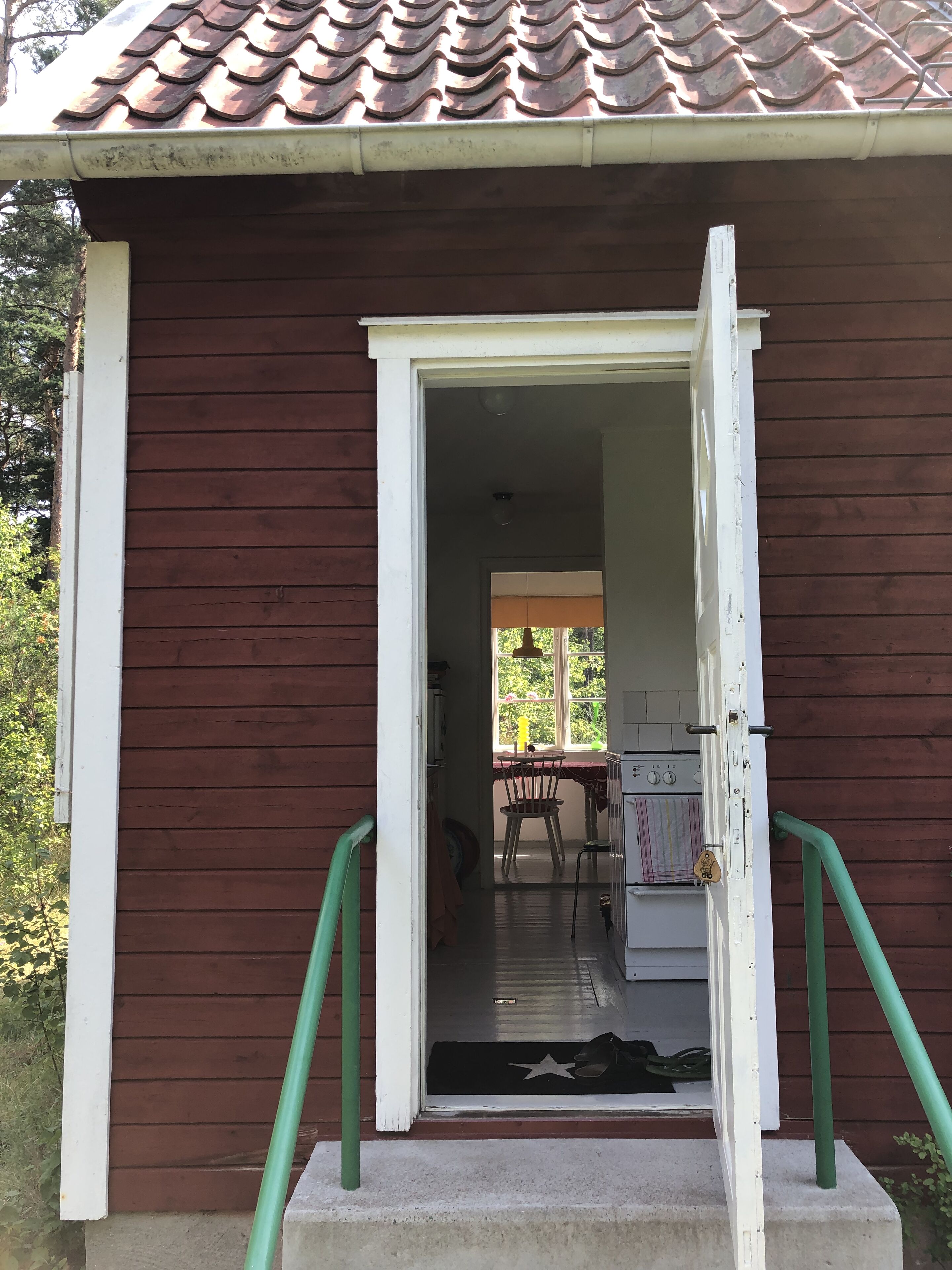 A wooden building with a brown exterior and a tiled roof. A door is open, revealing a kitchen area with a stove, a chair, and a table.