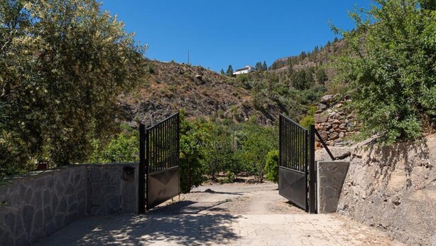 A gated entrance with a stone wall on one side and a dirt path leading to a hill.