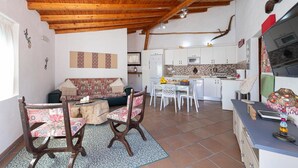 A cozy living area with a wooden ceiling, a sofa, a dining table with chairs, and a kitchen in the background.