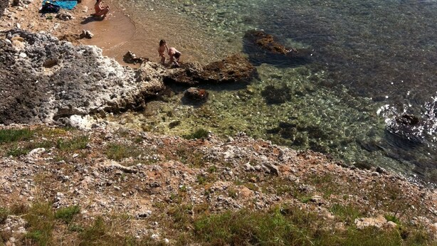 Une plage de rochers avec une eau claire, des gens qui prennent un bain de soleil, et une colline boisée en arrière-plan.