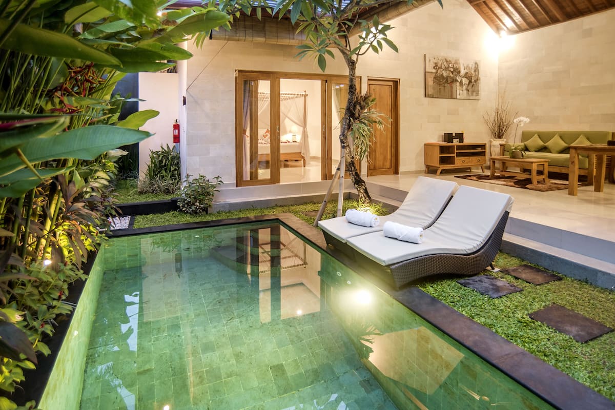 A modern outdoor pool area with a glass enclosure, surrounded by lush greenery and a cozy seating area with a sofa and lounge chairs.