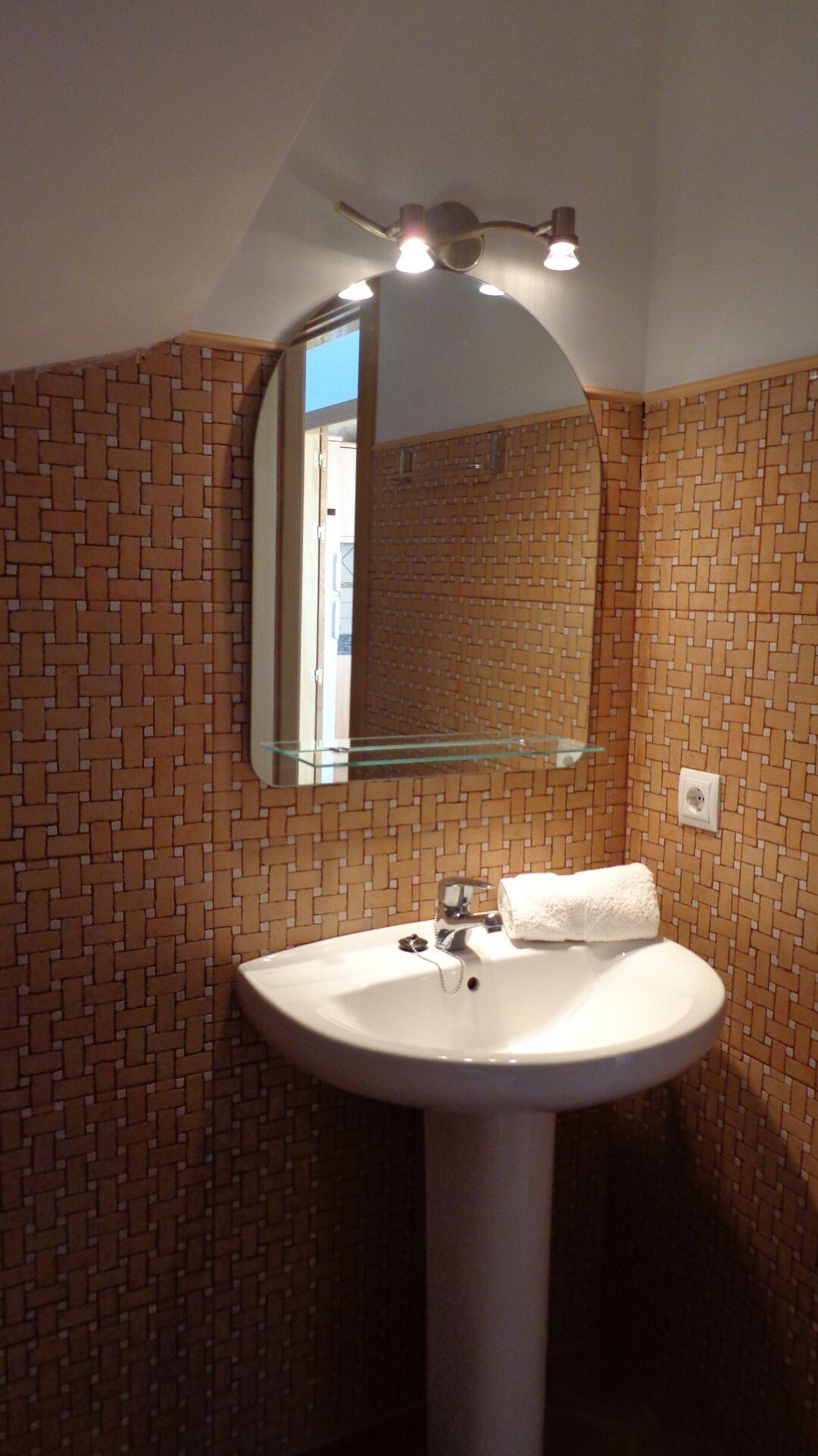 Un baño con un lavabo blanco, un estante de vidrio, un espejo y una lámpara sobre el espejo.