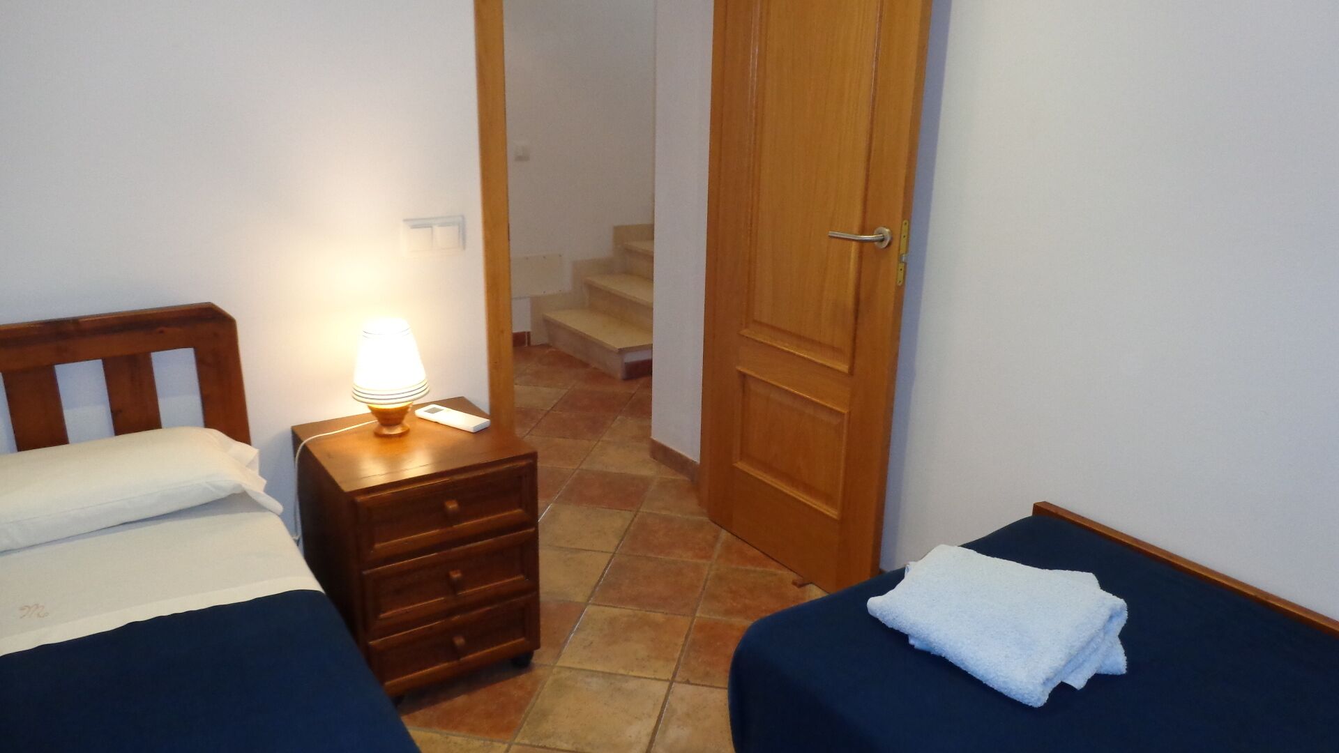 A bedroom with two single beds, a nightstand with a lamp, a wooden door, and a staircase in the background.