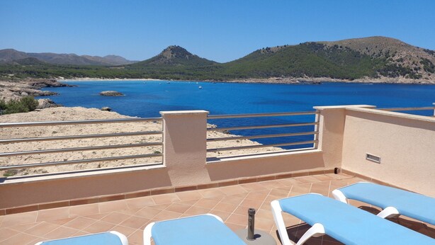 Een terras met blauwe stoelen met uitzicht op een kustgebied met helderblauw water en heuvels op de achtergrond.