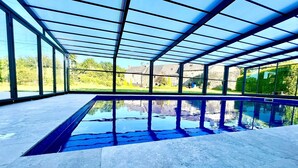 Une piscine dotée d’une enceinte en verre, entourée d’une terrasse en pierre et de grandes fenêtres offrant une vue sur la verdure et les bâtiments.