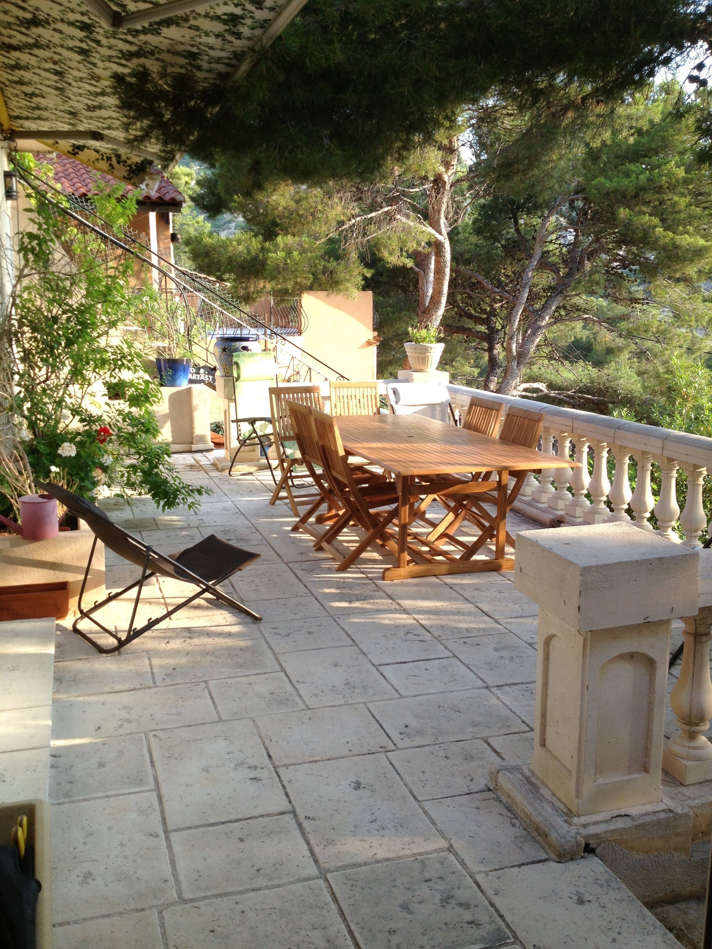 An outdoor patio with a wooden table and chairs, surrounded by potted plants and a stone balustrade.