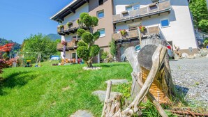 A multi-story building with balconies, a grassy area, and a wooden sculpture. - Nice apartment for 4 guests with WIFI, TV and balcony (Pians)