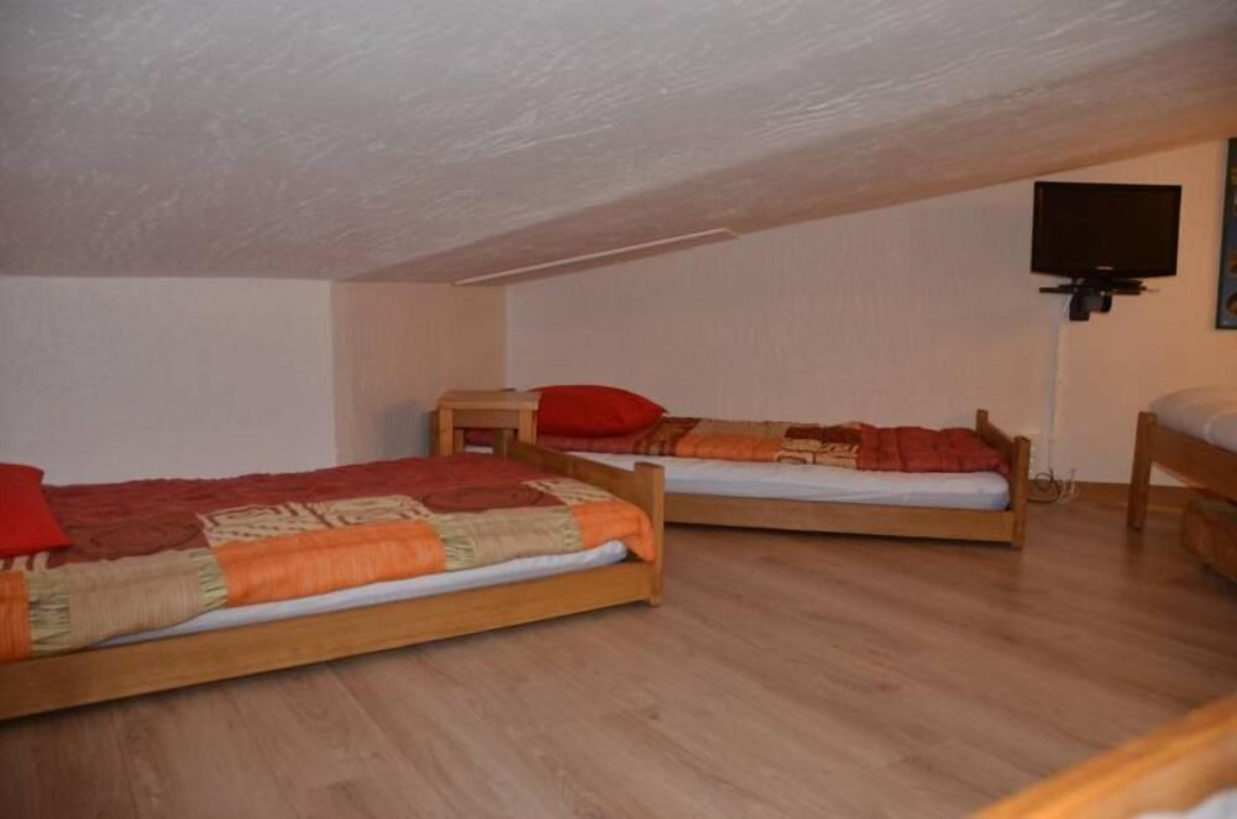A room with two wooden beds, each with a red pillow and a patterned blanket. A television is mounted on the wall.
