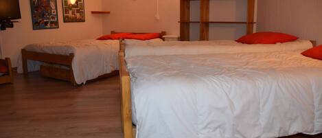 A room with two wooden beds, each with white bedding and red pillows. There is a wooden shelf unit against the wall and a wall-mounted television.