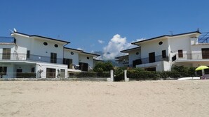 A sandy beach area with two white buildings with balconies, a clear blue sky, and a few clouds.