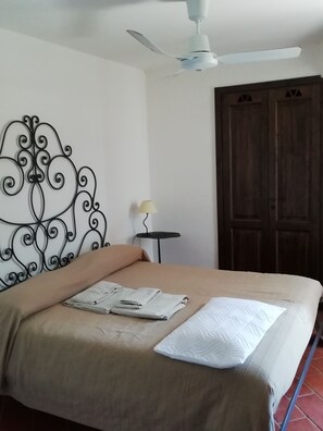 A bedroom with a bed, a ceiling fan, a decorative wall piece, a small table with a lamp, and a wooden door.