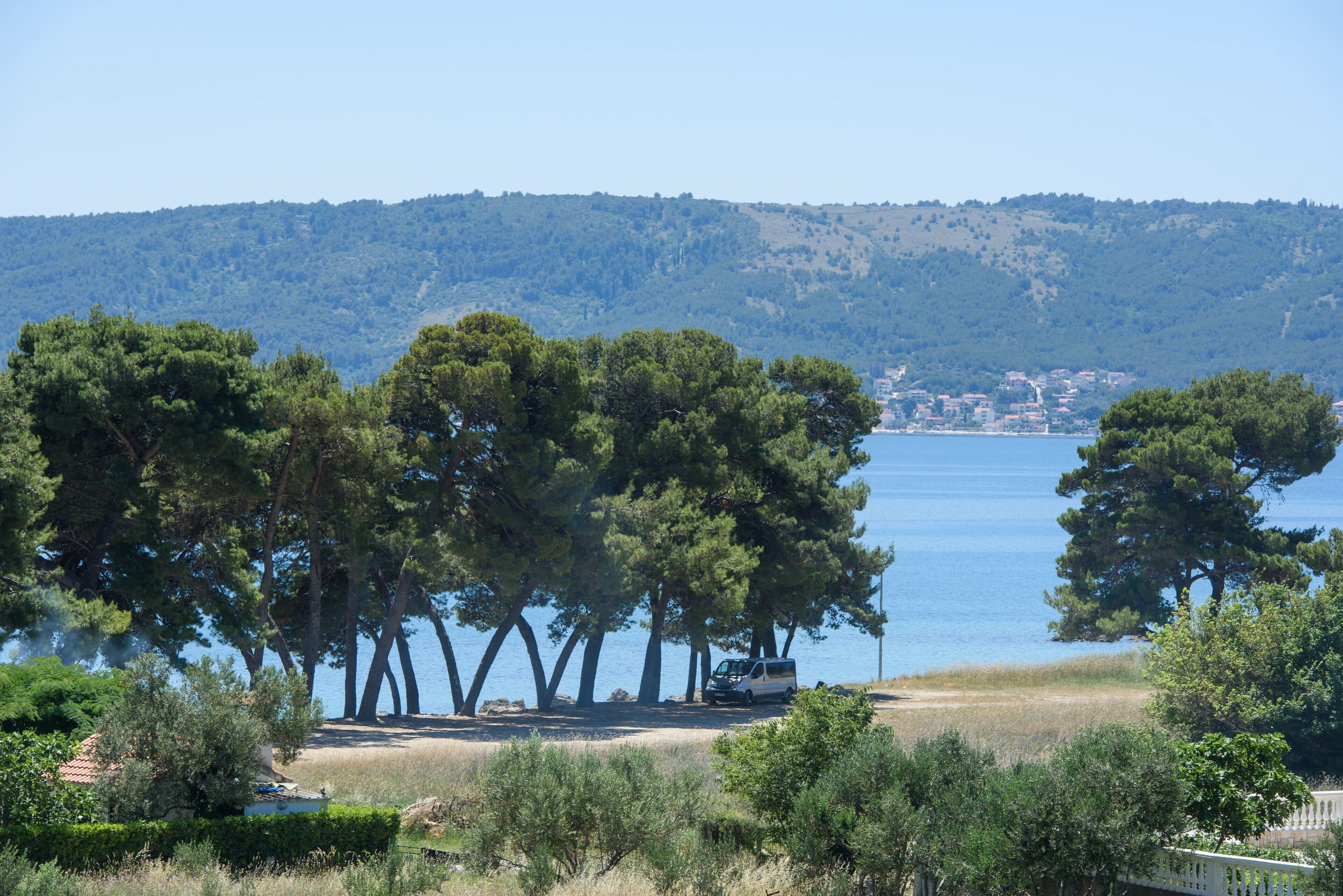 Une zone côtière où un fourgon est garé près d’une plage, entouré d’arbres, avec une vue lointaine sur une ville.