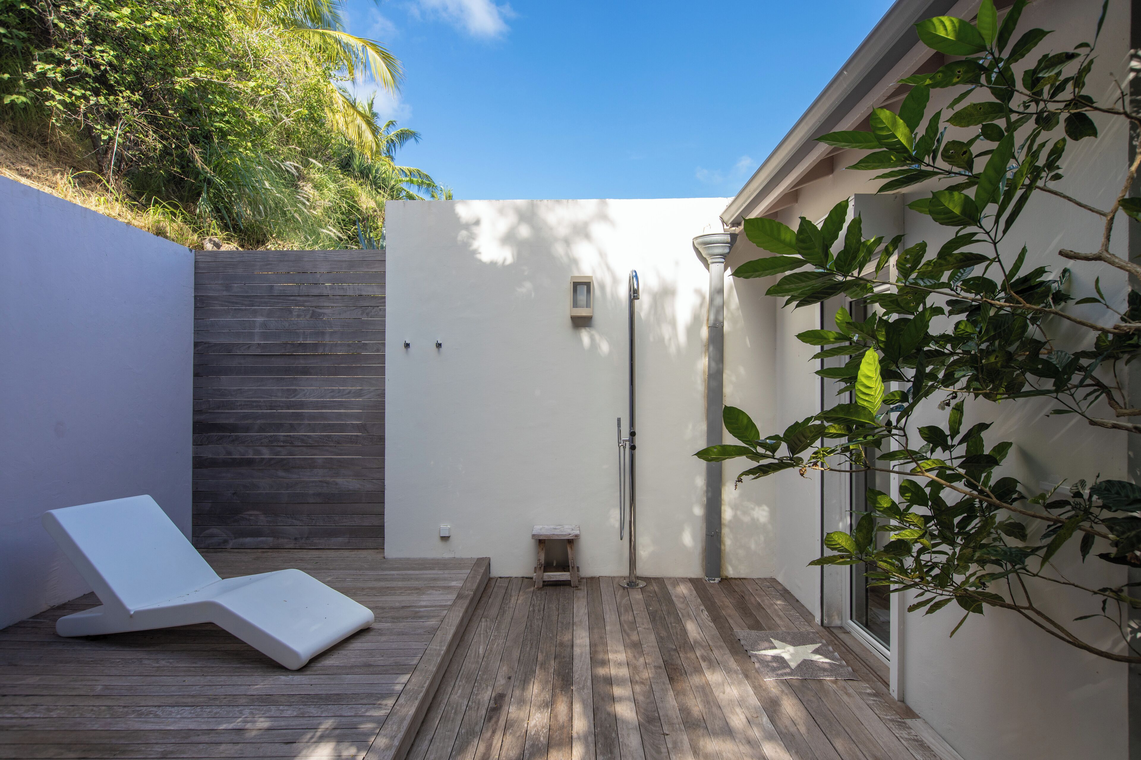 A wooden deck with a white lounge chair, a small wooden stool, and a shower area with a glass door.