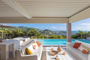 Zona de piscina con mobiliario blanco, vista al mar y montañas de fondo.