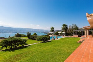 Zona de piscina con vista al mar, rodeada de vegetación y un patio empavedado.
