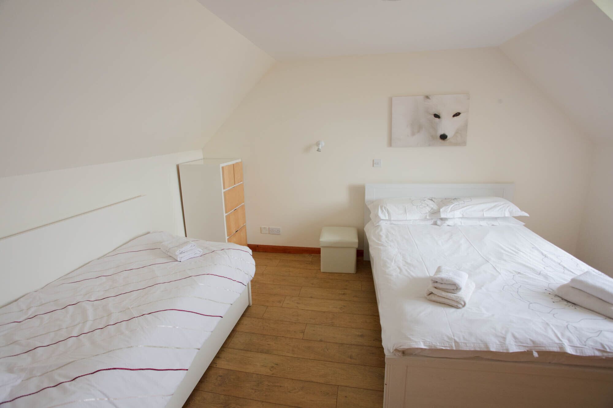 A small, clean room with two single beds, a wooden floor, and a white wall with a framed picture.