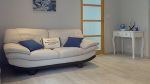 A living room with a beige sofa, two blue cushions, a small white side table with a mirror and a vase, and a painting of a hammock on a wall.