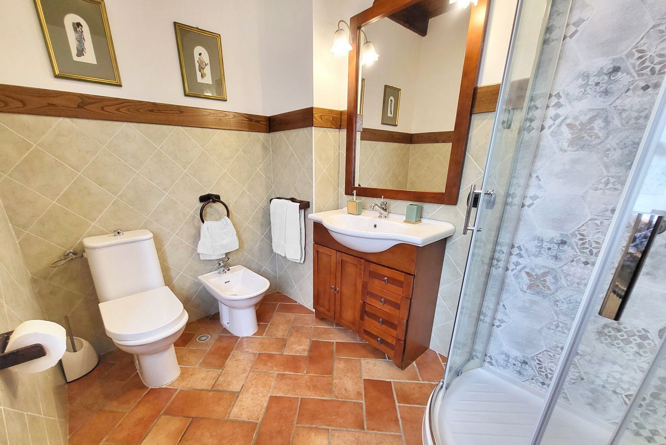 A bathroom with a toilet, bidet, sink, wooden vanity, and a shower area.