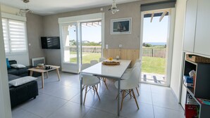 A modern living room with a sofa, a coffee table, and a TV. There is a dining area with a table and chairs. The room has large windows and a view of the outdoors.