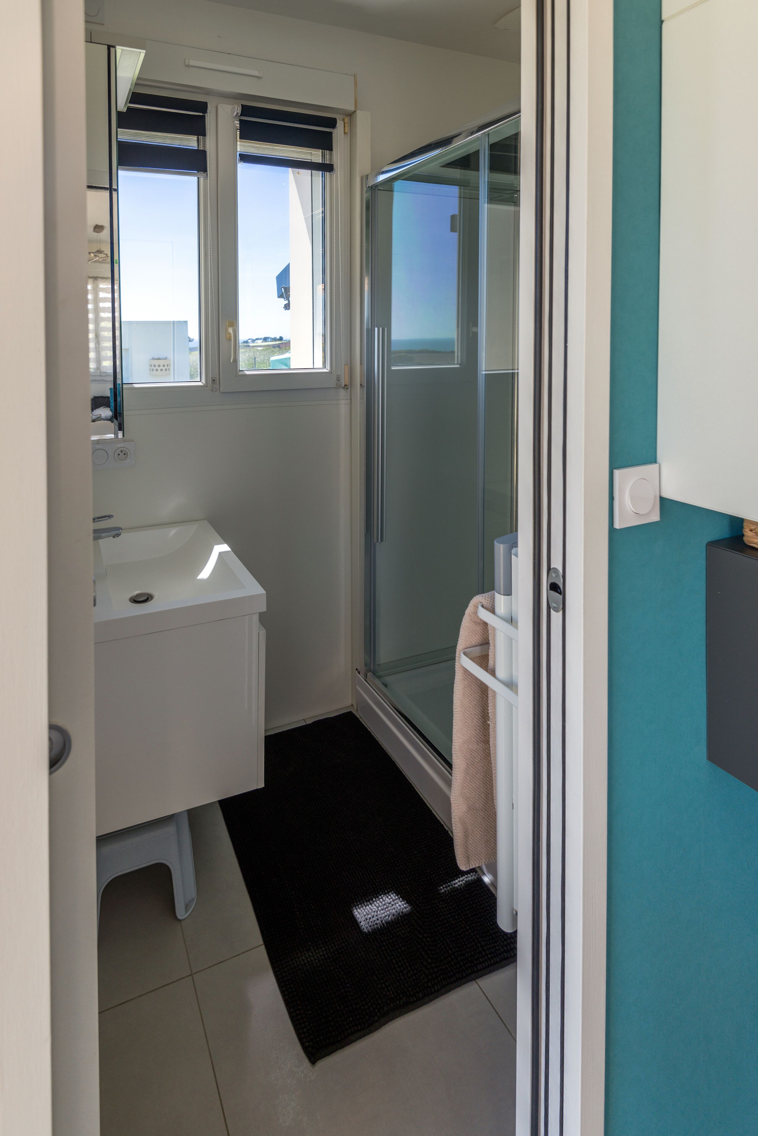 Ein kleines Badezimmer mit Waschbecken in Weiß, einer Duschkabine aus Glas und einer schwarzen Matte im Eingangsbereich.