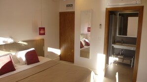 Une chambre d’hôtel comprenant un lit, une tête de lit en bois, un miroir, une salle de bain avec un lavabo et un miroir, et un panneau indiquant la sortie.