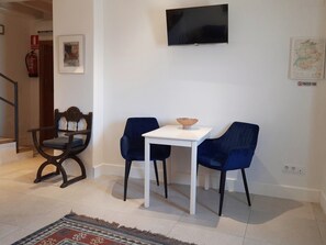 Un salon avec une table blanche, deux chaises bleues, une chaise en bois, un téléviseur fixé au mur, un extincteur et un tapis.