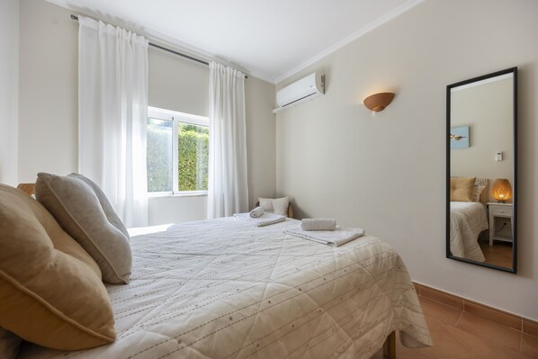 Een hotelkamer met een bed, een raam met gordijnen, een airconditioningapparaat, een wandlamp en een spiegel.