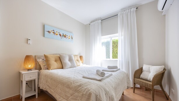 Un dormitorio con una cama, una mesita de noche, una lámpara, una silla y una ventana con cortinas.