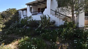 Un bâtiment blanc avec un toit recouvert de tuiles, entouré d’un jardin où poussent différentes plantes et fleurs.