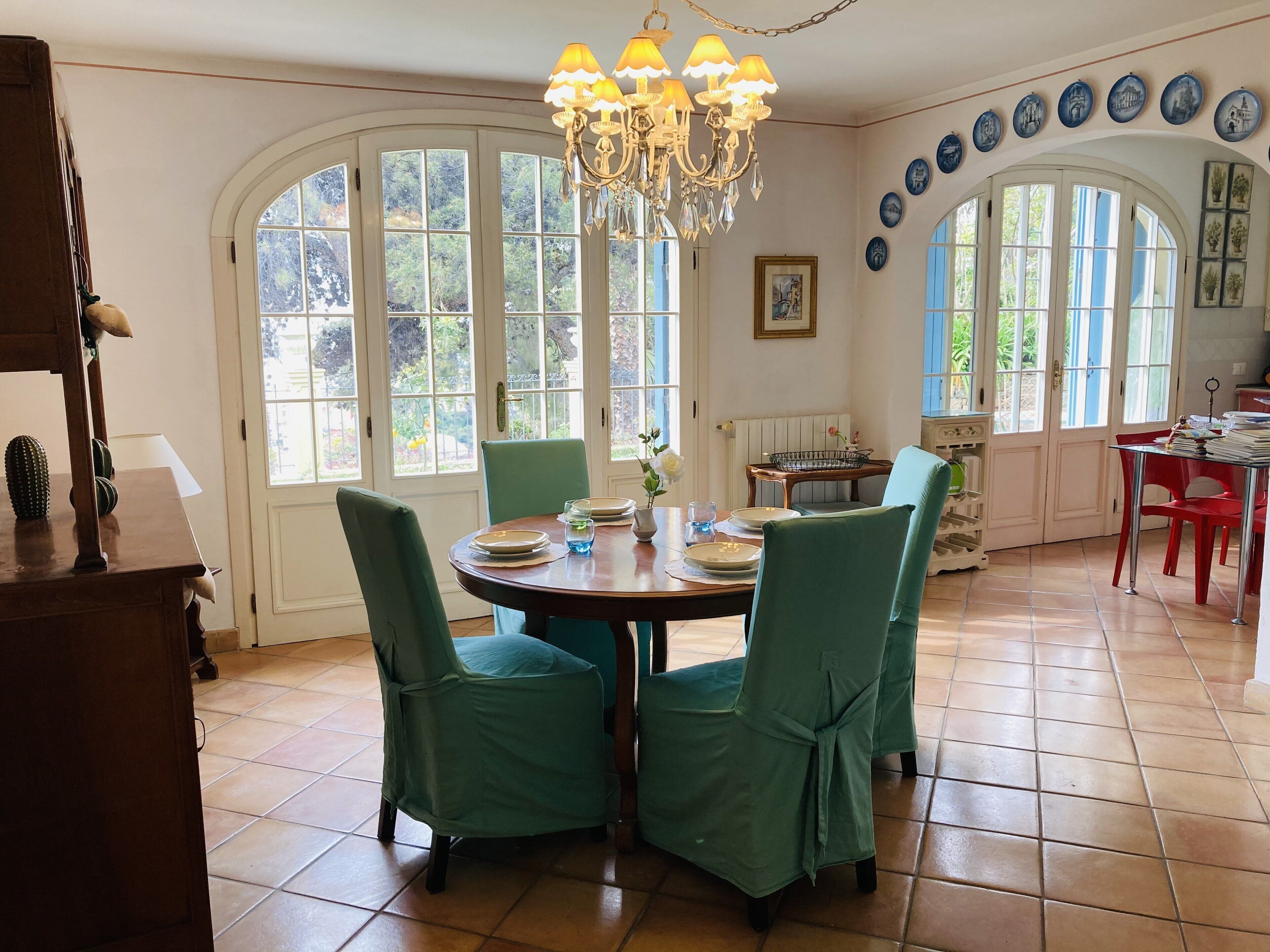 A dining area with a round table, green chairs, and a chandelier. There are large windows and decorative plates on the wall.