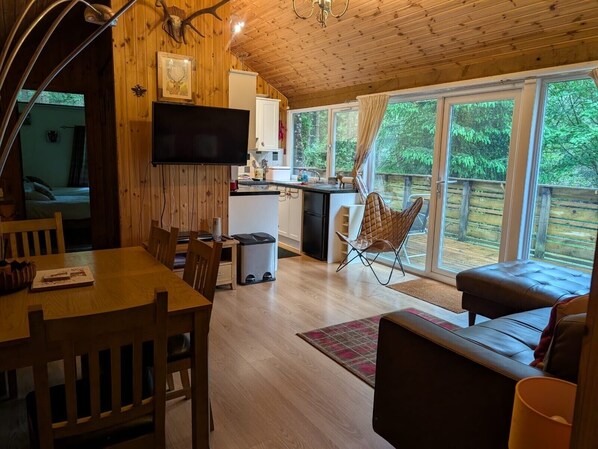 A wooden cabin interior with a dining area, kitchen, and a large sliding glass door leading to an outdoor deck.