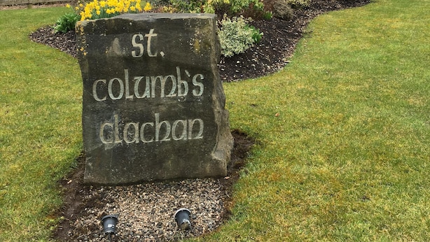 Un letrero de piedra con la inscripción "St. Columb's Clachan" en un área ajardinada con césped, arbustos y una valla de madera al fondo.