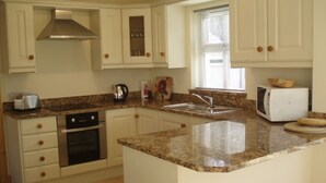 Una cocina con armarios blancos, encimera de granito, campana extractora de acero inoxidable, horno empotrado, microondas y una ventana con persianas.