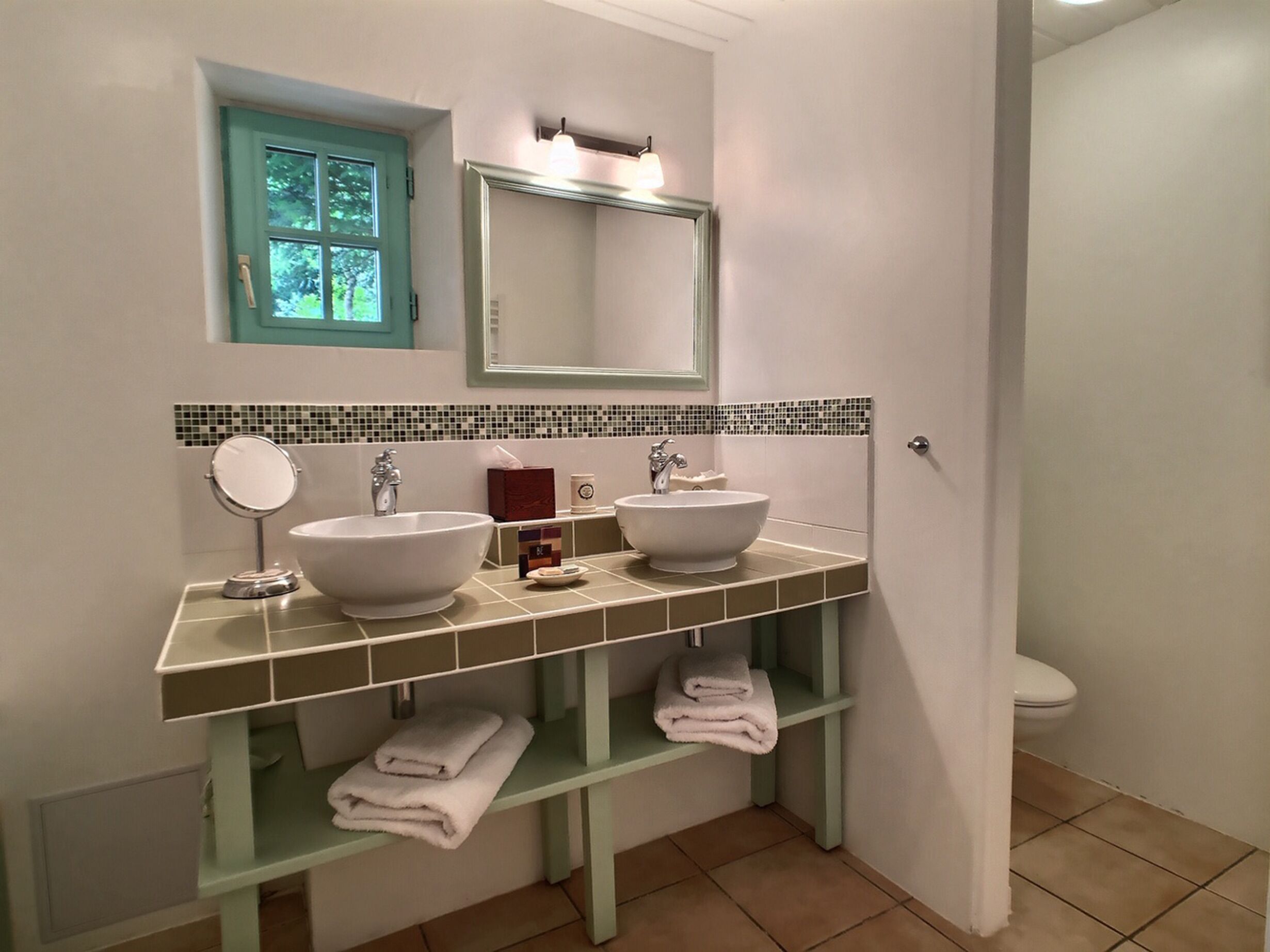 Une salle de bain avec deux lavabos, un miroir et des toilettes. Il y a des serviettes sur les étagères et une fenêtre donnant sur les arbres.