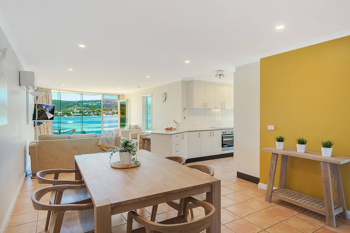 A modern kitchen with white cabinets, a wooden dining table, and a view of the sea through large windows.