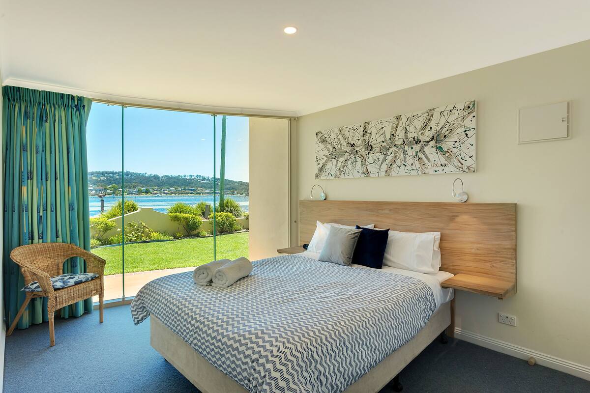 A hotel room with a large bed, a view of the sea, a wicker chair, and a painting on the wall.