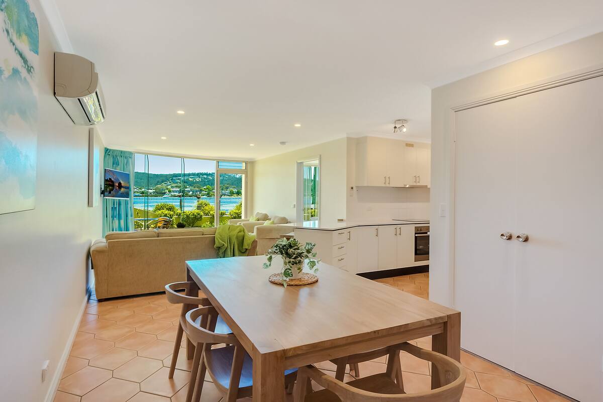 A modern kitchen with white cabinets, a dining area with a wooden table and chairs, and a view of a coastal landscape through large windows.