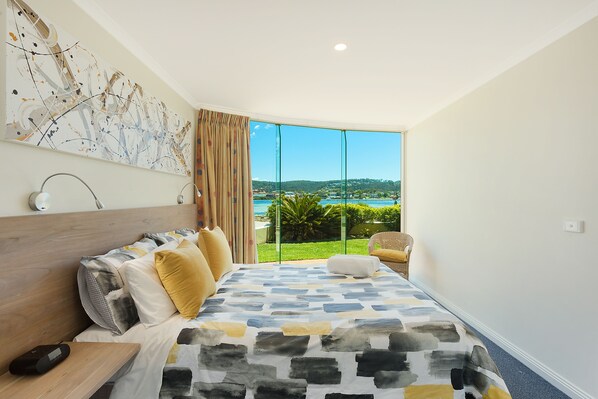 A bedroom with a large bed, a bedside table, a lamp, a painting on the wall, and a view of the outdoors. - Cetacea Luxury Apartments (Merimbula)