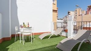 Eine Dachterrasse mit Kunstrasen, weißen Stühlen und einem Tisch mit Getränken und Früchten.