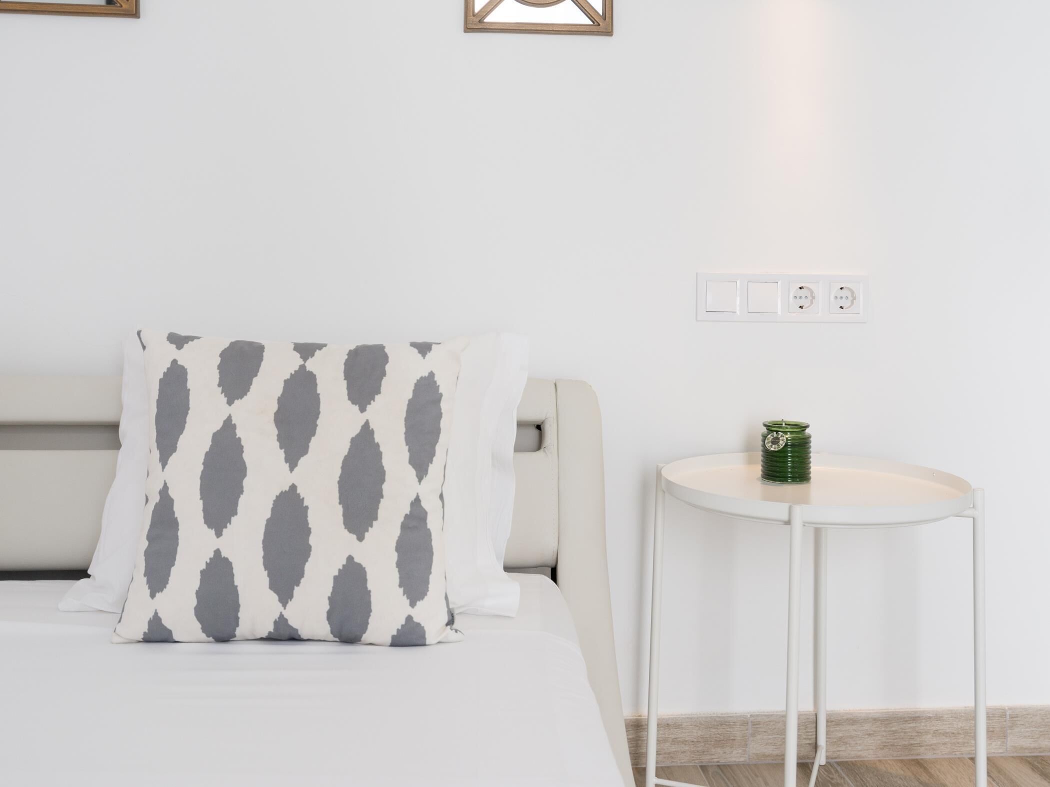A neatly arranged bedroom with a bed, a pillow with a geometric pattern, a bedside table with a green container, and a wall socket.