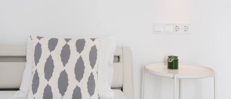 A neatly arranged bedroom with a bed, a pillow with a geometric pattern, a bedside table with a green container, and a wall socket.