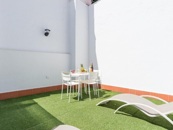 Un patio con una mesa y sillas blancas, un frutero pequeño y una botella de vino.