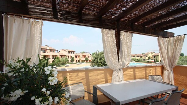 A covered outdoor seating area with a table and chairs, overlooking a pool and buildings.