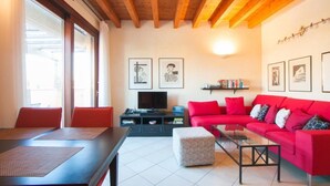 A modern living room with a red sofa, a glass coffee table, and a television on a black stand. There are framed pictures on the wall and a bookshelf with books.