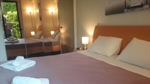 Een opgemaakt bed met witte lakens en kussens, een nachtkastje met een lamp, en een spiegel die de kamer weerspiegelt.