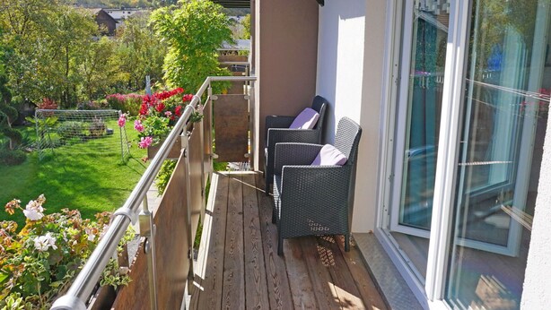 A balcony with wicker furniture, a glass door, and a view of a garden and houses in the background.
