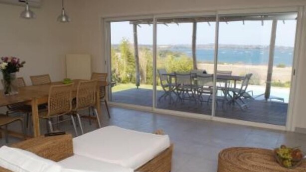 Une chambre moderne dotée d’une grande porte-fenêtre coulissante qui donne sur un patio extérieur. L’intérieur comprend un canapé blanc, une table basse en osier et un coin repas avec des chaises en osier.
