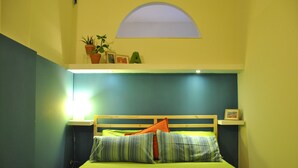 A single bed with a wooden headboard, green bedding, and two pillows. There is a small shelf with a potted plant and a framed picture above the bed.