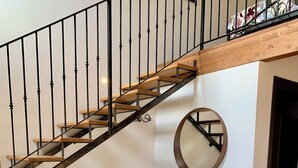 Una escalera con escalones de madera y barandillas negras, un televisor sobre un soporte de madera, un espejo redondo en la pared y un radiador.